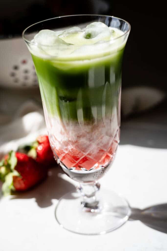 Layered iced strawberry matcha latte in a tall crystal glass, with red strawberry, white milk, and green matcha layers, surrounded by fresh strawberries.