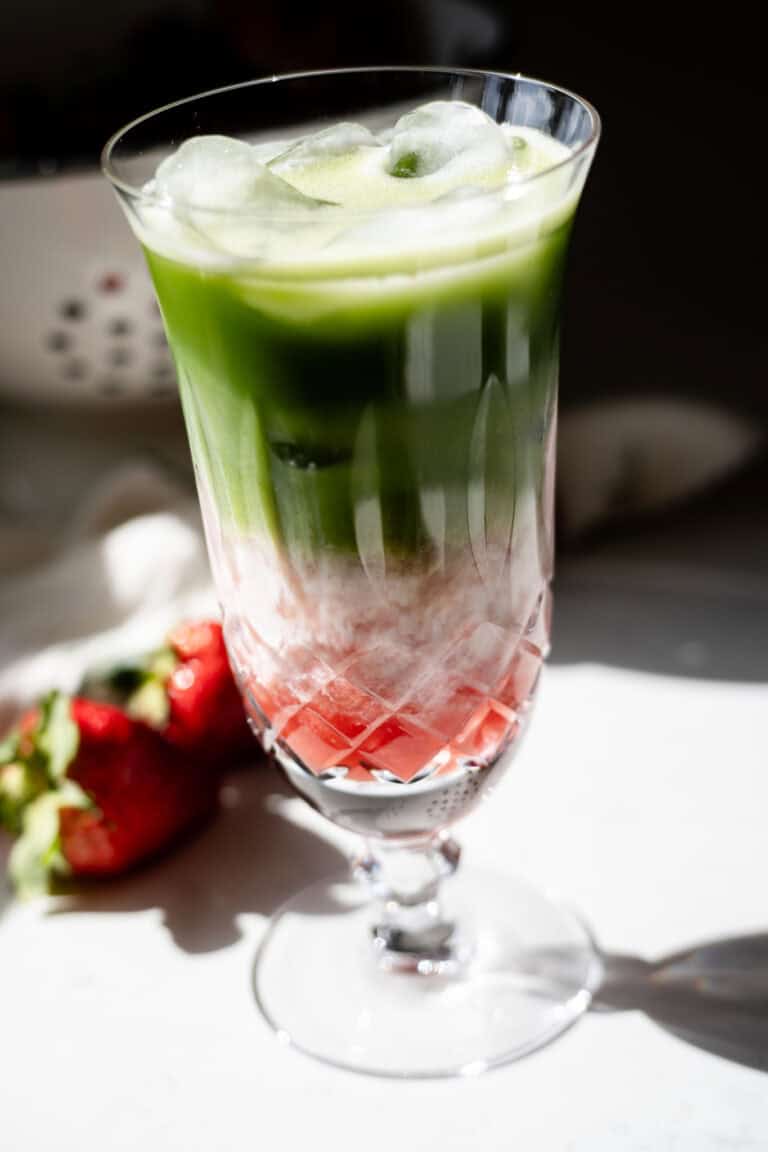 Layered iced strawberry matcha latte in a tall crystal glass, with red strawberry, white milk, and green matcha layers, surrounded by fresh strawberries.