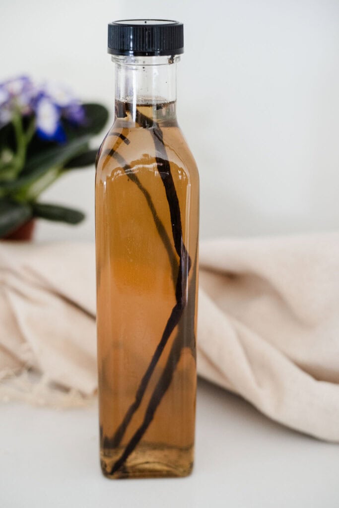 A clear glass bottle filled with homemade vanilla syrup, set against a background of greenery and beige.