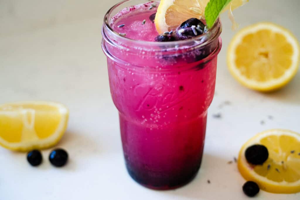 A glass of blueberry lavender lemonade filled with ice, garnished with fresh blueberries, a lemon slice, and a sprig of lavender.