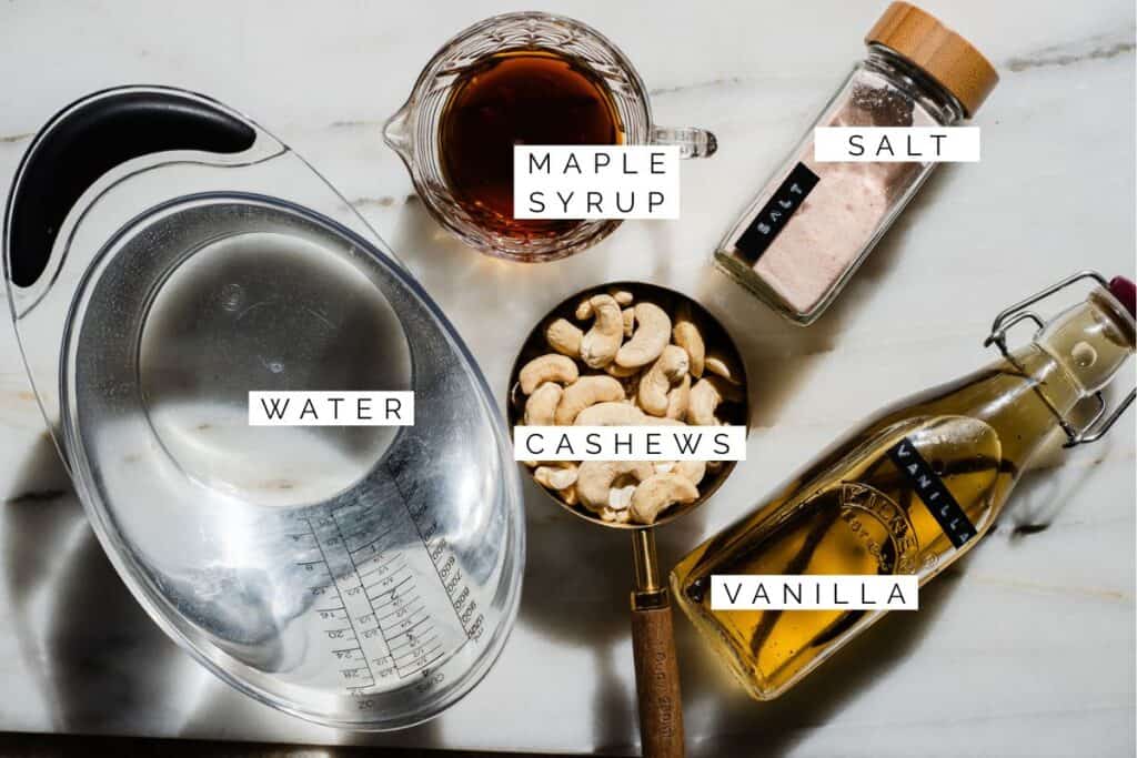 A simple, clean flat lay of the ingredients for homemade cashew milk: raw cashews, maple syrup, vanilla extract, and a pinch of sea salt