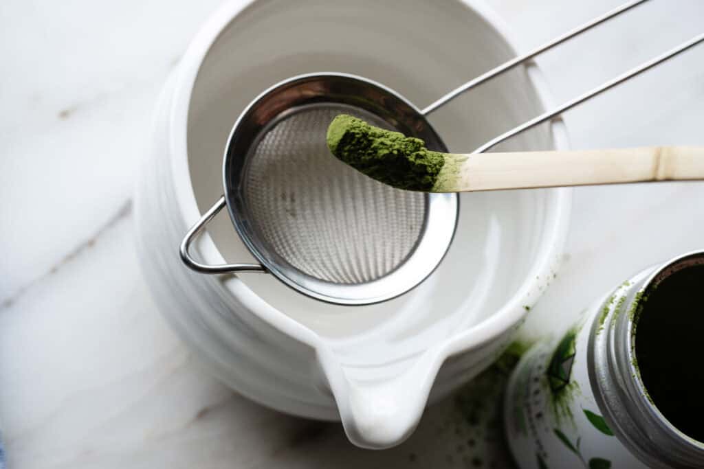 white bowl, stainless sifter and wooden scoop with green matcha powder